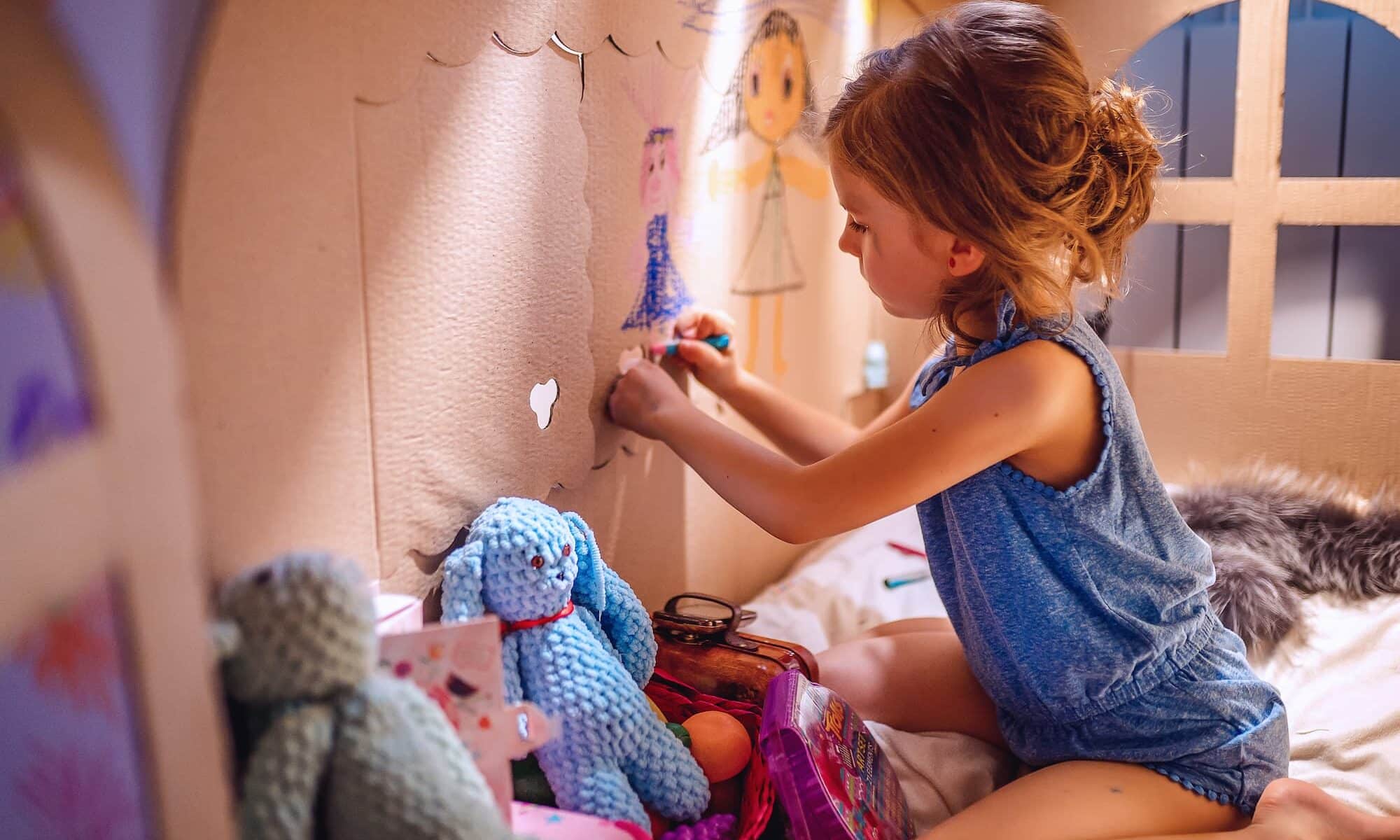 Vue latérale d'une fille dessinant avec du crayon sur le mur d'une maison en carton assise à l'intérieur et s'amusant à la maison.