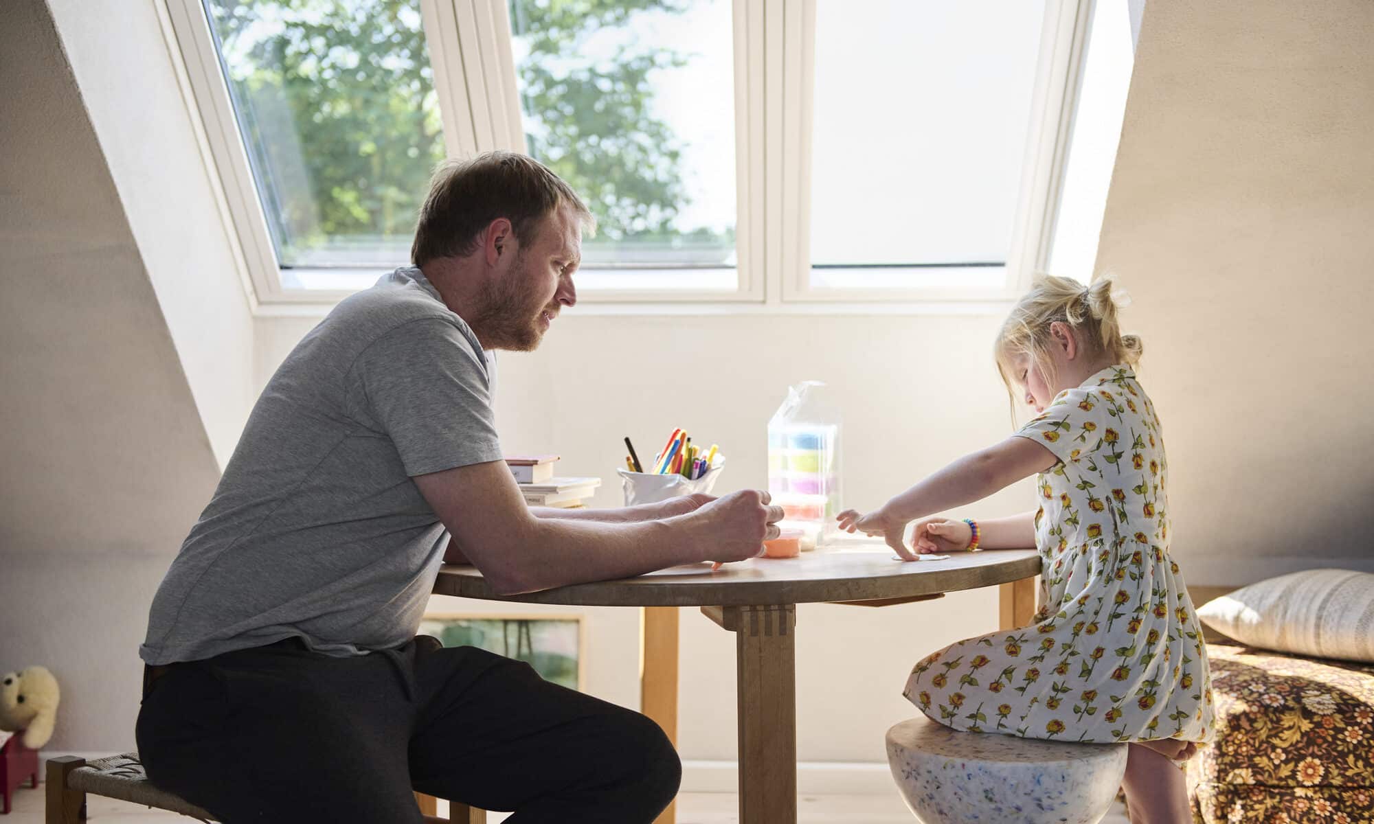 Salle de jeux mansardée avec plafond en pente et grande fenêtre de toit, enfant et adulte assis à table.