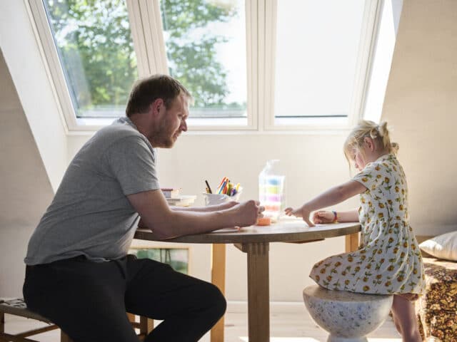 Salle de jeux mansardée avec plafond en pente et grande fenêtre de toit, enfant et adulte assis à table.