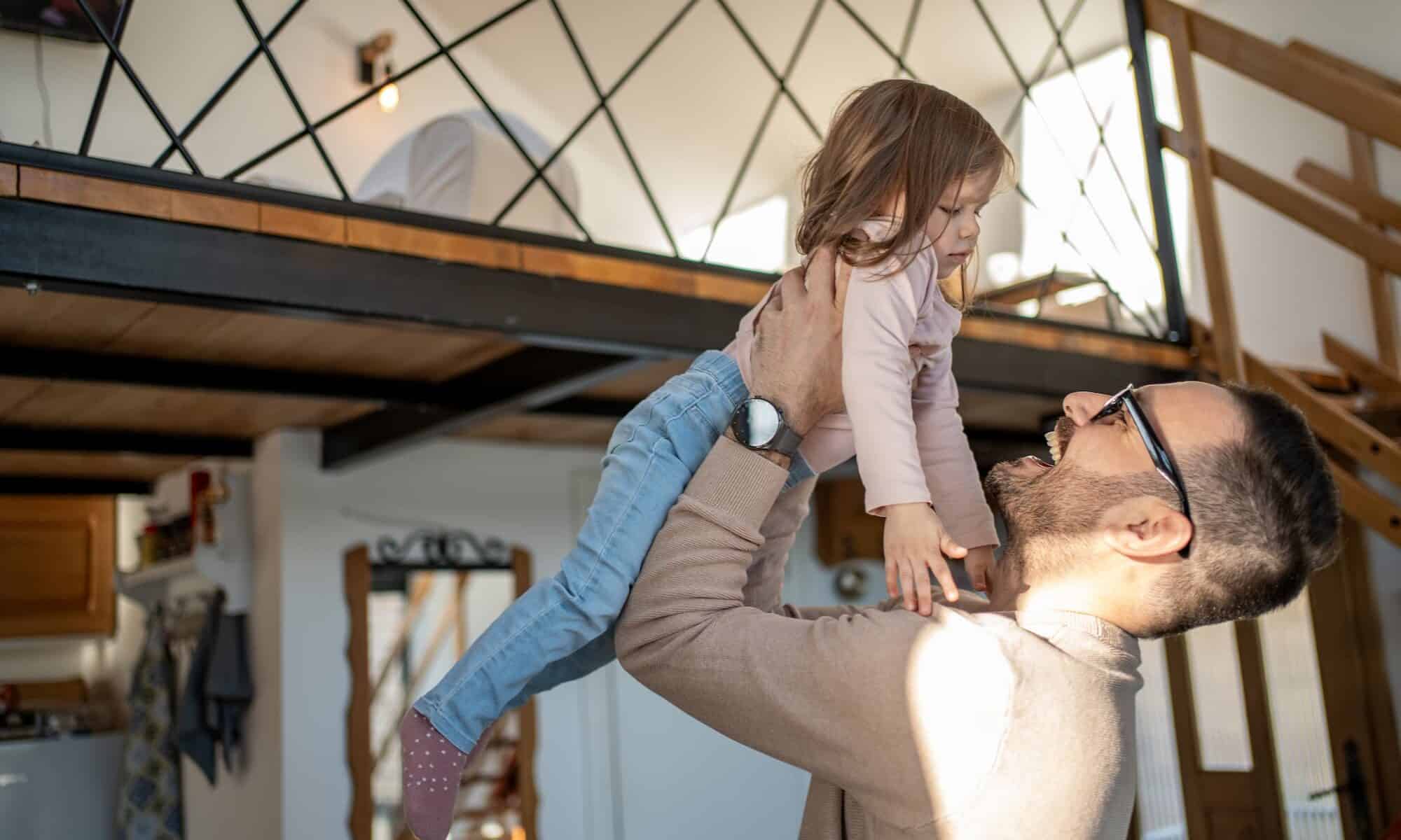 Heureux père soulevant sa fille dans les airs dans leur appartement en duplex, avec une mezzanine