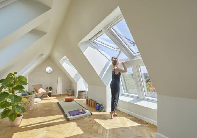 Salle de yoga mansardée avec plafond en pente et fenêtres de toit VELUX, femme ajustant la fenêtre.