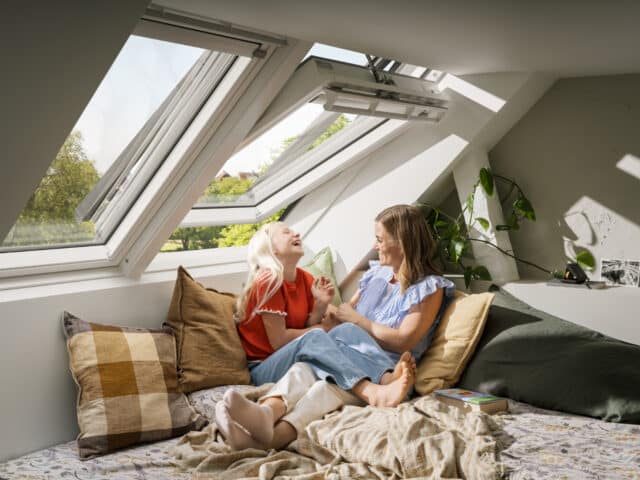 Chambre mansardée avec plafond incliné et fenêtre de toit VELUX ouverte, deux personnes se reposant sur le lit.
