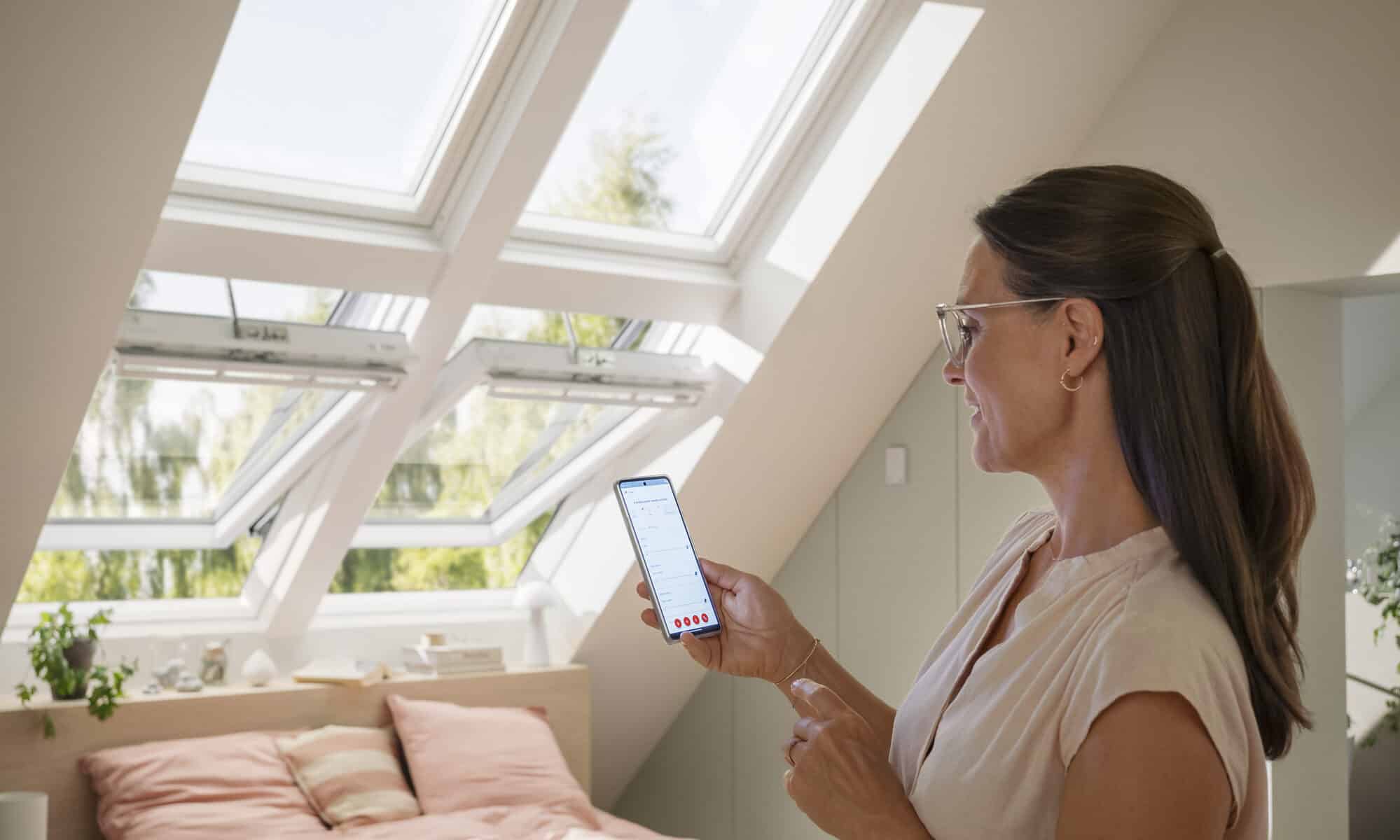 Chambre mansardée avec fenêtres de toit VELUX ouvertes, lumière naturelle et vue sur la verdure.