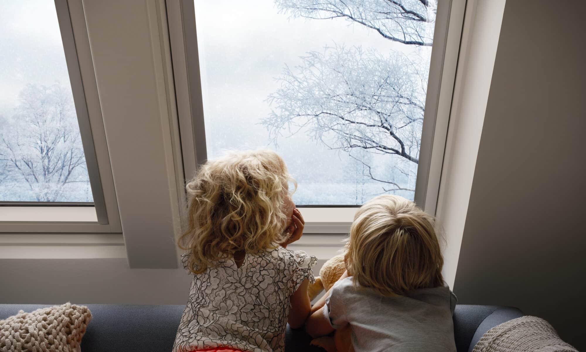 Des enfants assis sur un canapé, regardant par une fenêtre de toit VELUX les arbres enneigés.