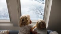 Des enfants assis sur un canapé, regardant par une fenêtre de toit VELUX les arbres enneigés.