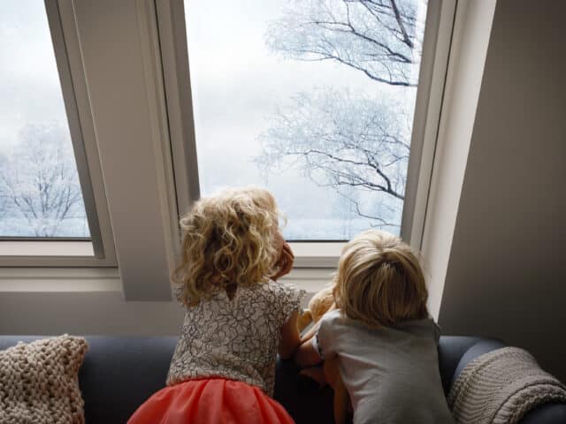 Des enfants assis sur un canapé, regardant par une fenêtre de toit VELUX les arbres enneigés.