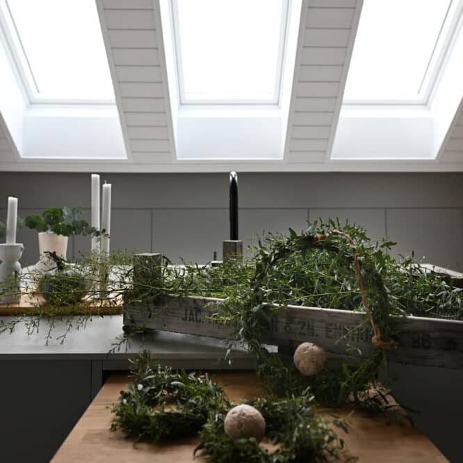 Décorations de Noël faites de couronnes de feuilles, chandeliers et bougies dans les tons verts et beige, posés sur le rebord d'une fenêtre de toit sous les combles.