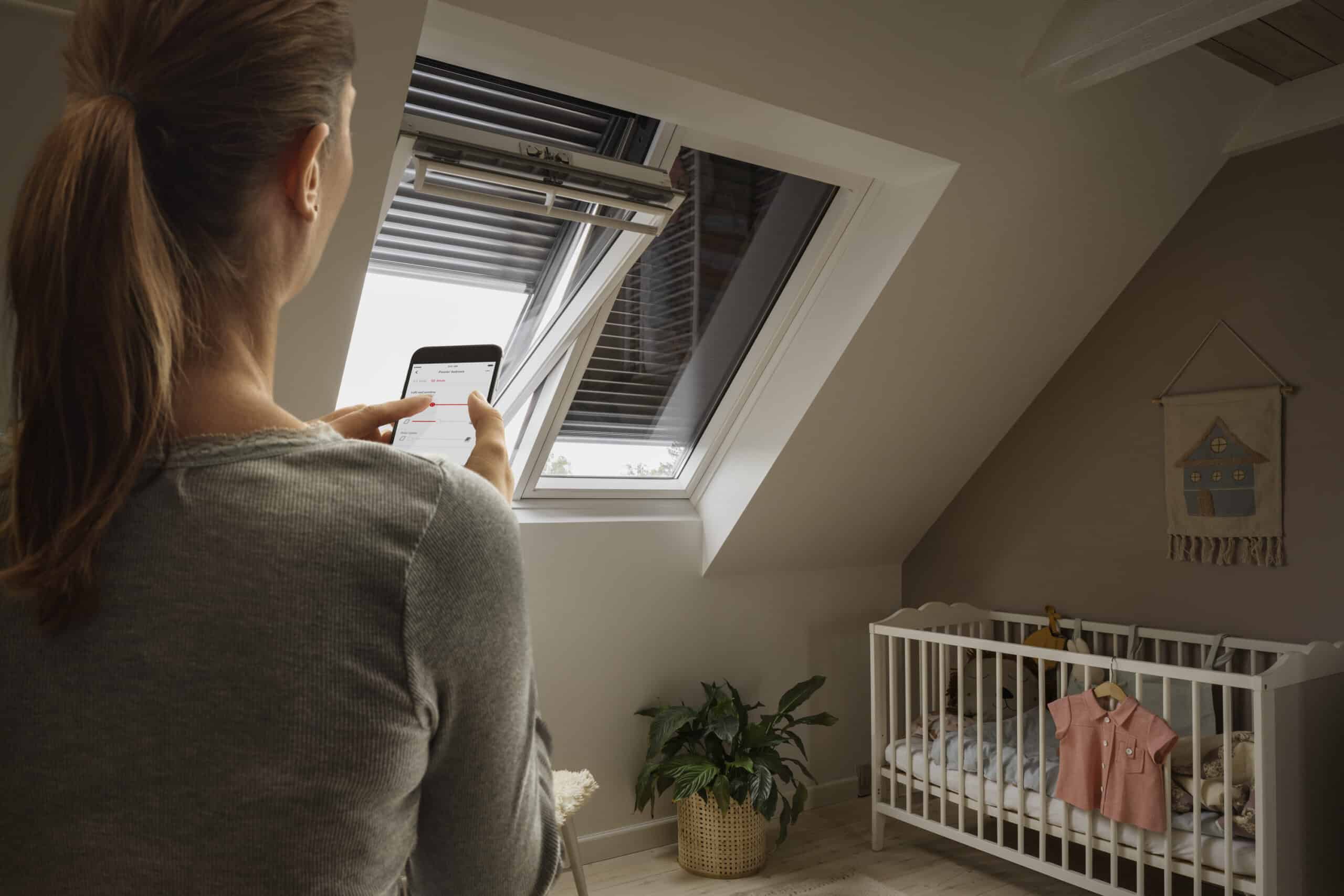 Chambre d'enfant avec fen&ecirc;tre de toit VELUX ouverte, une femme utilise un smartphone pour la contr&ocirc;ler.