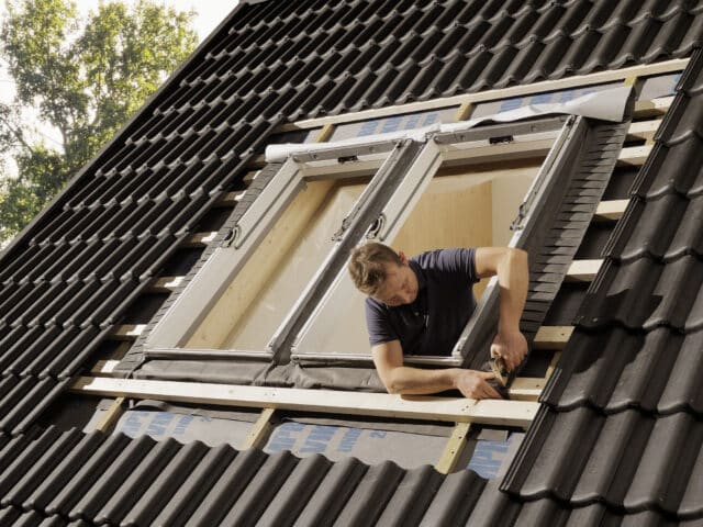 Ouvrier installant une fenêtre de toit VELUX sur un toit en tuiles noires à forte pente.