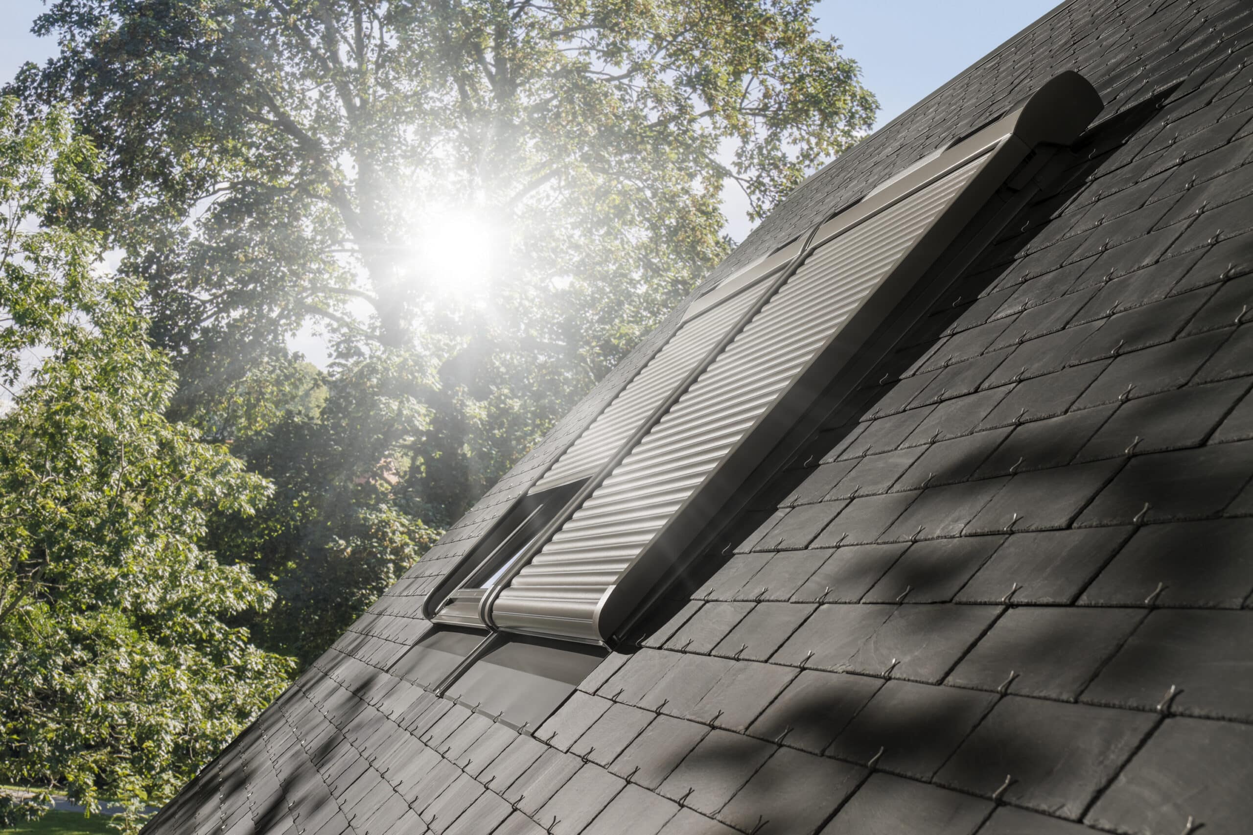 Toit en ardoise avec fen&ecirc;tres VELUX ferm&eacute;es et volet roulant solaire VELUX, la lumi&egrave;re du soleil filtrant &agrave; travers les arbres.