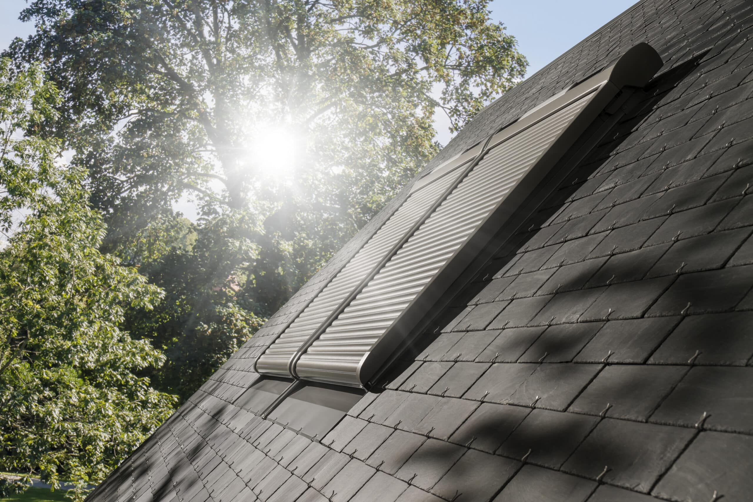 Toit en ardoise avec fenêtres VELUX fermées et volet roulant solaire VELUX, la lumière du soleil filtrant à travers les arbres.