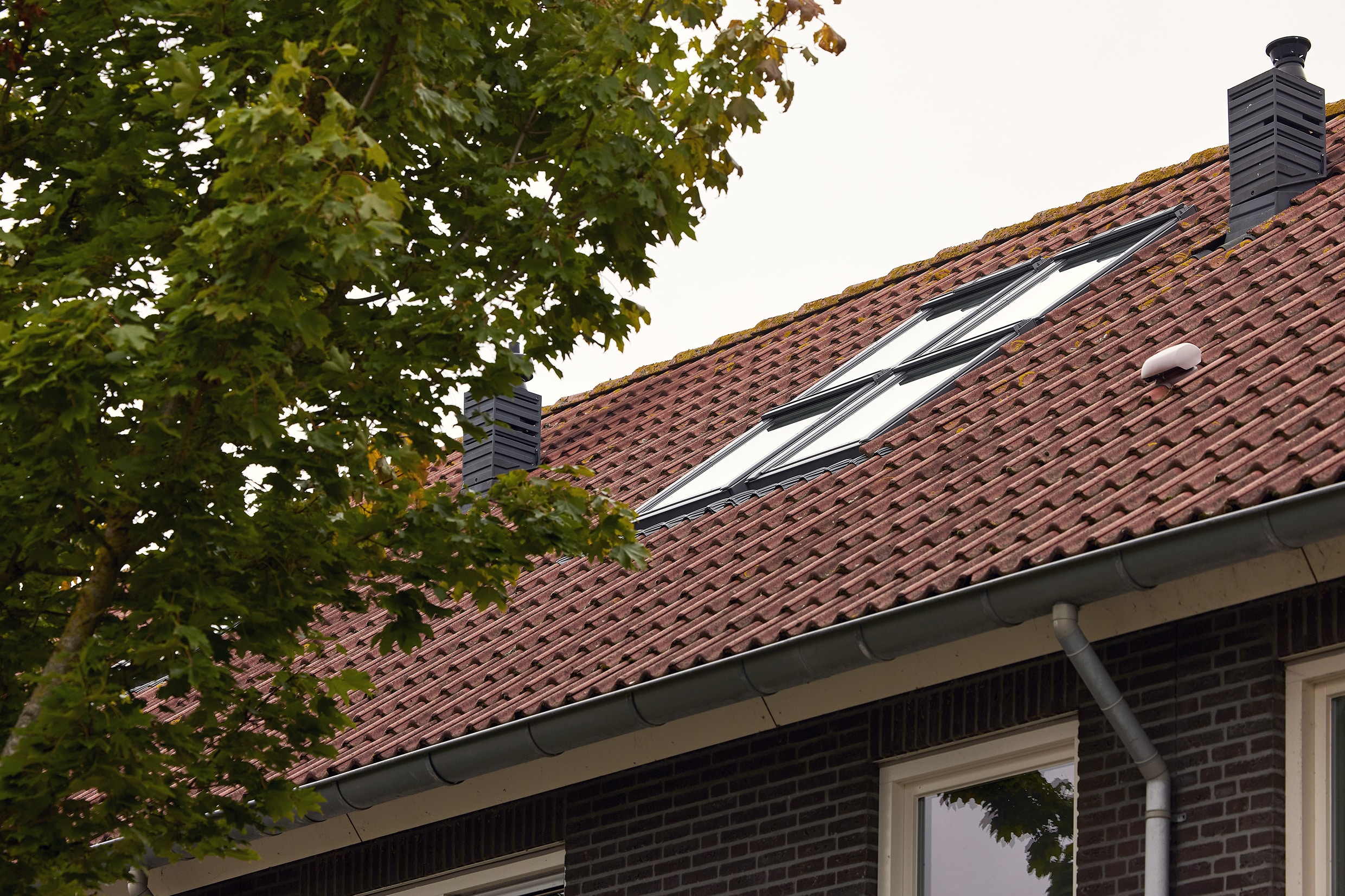 Toit en tuiles rouges avec fenêtre et cheminées en VELUX, entouré de verdure.