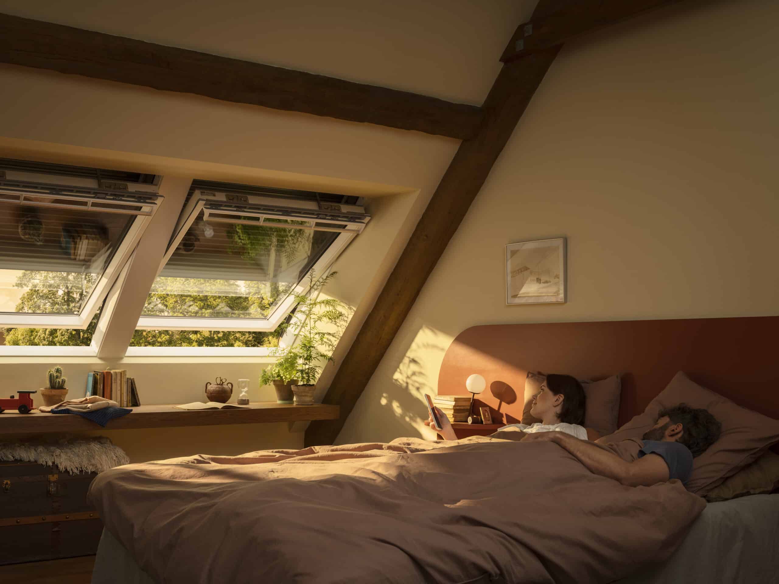 Chambre mansardée avec plafond en pente et fenêtres de toit VELUX ouvertes avec volet roulant solaire VELUX. Femme et homme au lit.