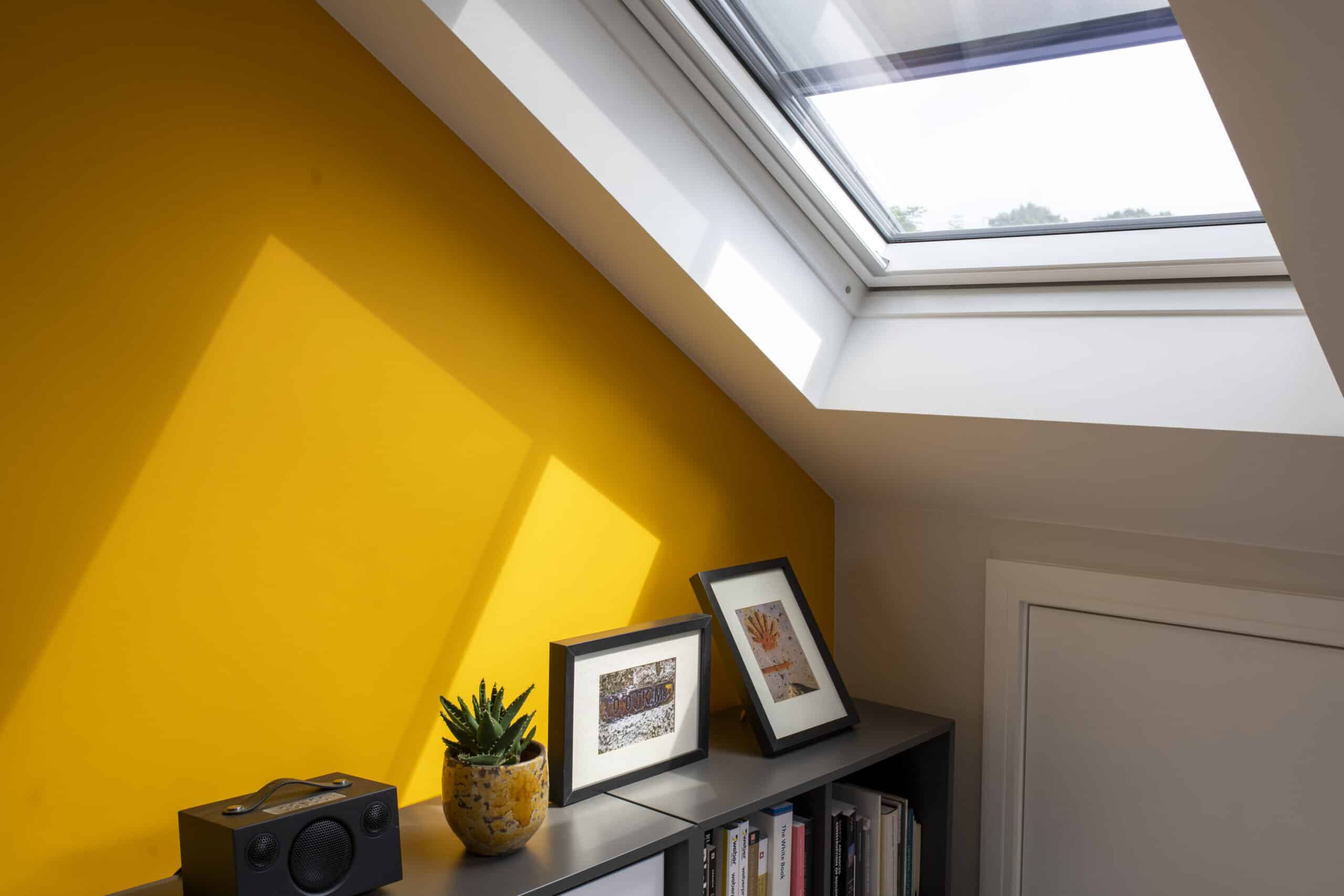 Chambre mansardée avec plafond en pente et fenêtre de toit VELUX, mur jaune et étagère.